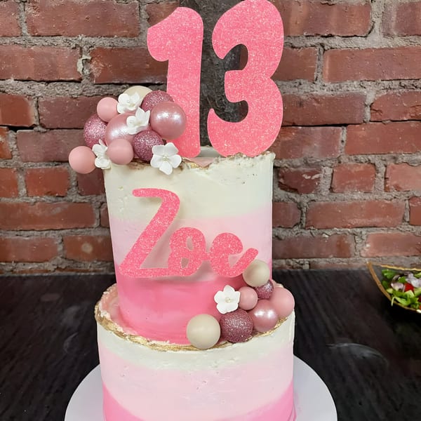 Two-tier pink ombre birthday cake with "Zoe" and "13" toppers, decorated with pink and white spheres.