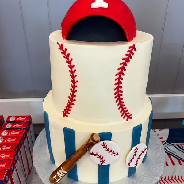 Two-tier baseball-themed cake with a red baseball cap, red stitching, blue stripes, and a bat.