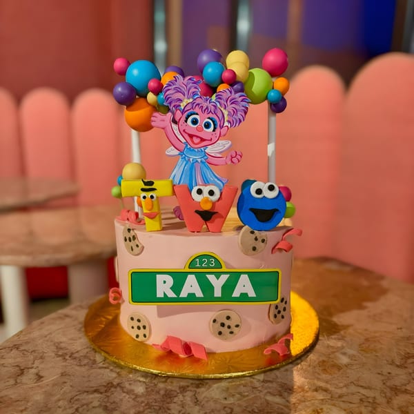 Pink birthday cake with Sesame Street characters, colorful balloons, and a "Raya" sign.