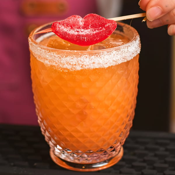 Orange cocktail in a textured glass with a sugar rim and a red candy lip garnish.